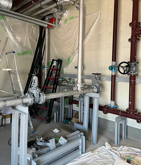 工事中の部屋に設置された金属製の配管とバルブ、周囲に脚立や工具、床にシートが広げられている様子。