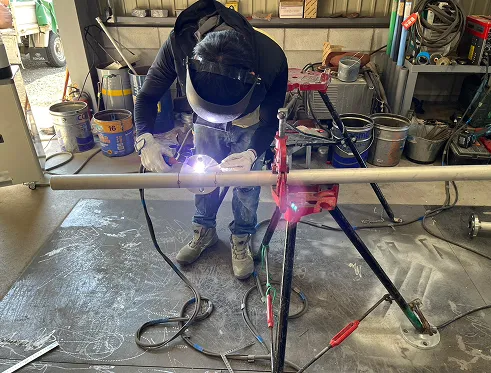 作業着を着た男性が溶接マスクをかぶり、工場内でパイプの溶接作業をしている。