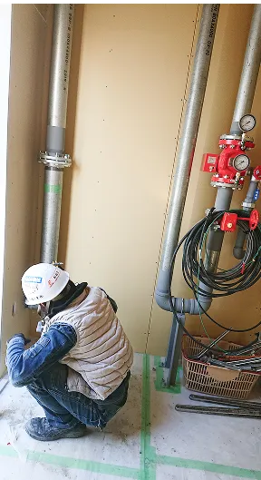 作業用ヘルメットとベストを着た作業者が室内で配管の点検作業をしている。