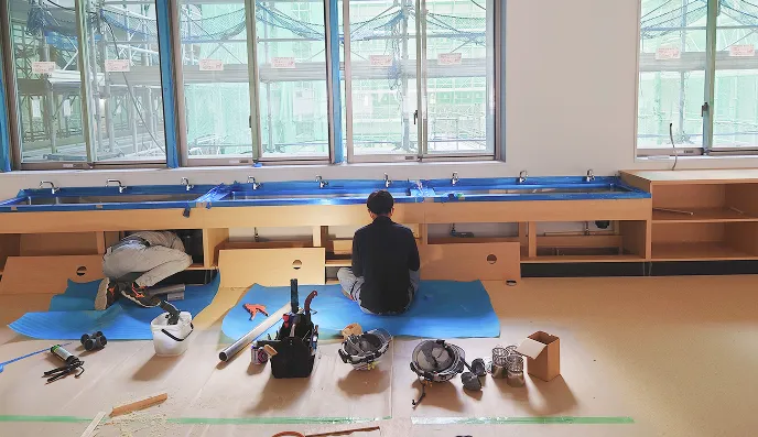 作業中の二人の人物が窓際の作業台の下に座り、工具や安全ヘルメットが床に並んでいる室内の建設現場。